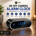 A black and silver alarm clock with a digital blue LED display, shown on a wooden table in a home setting with promotional text overlays highlighting its features like HD camera, night vision, motion detection, and remote monitoring.